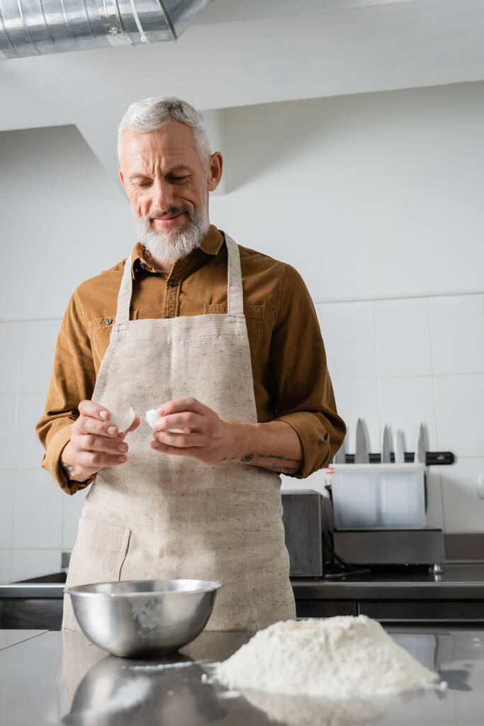 Lächelnder älterer Koch mit Eierschale in der Nähe von Mehl und Schüssel auf dem Tisch, während er in der Küche kocht  - Foto, Bild