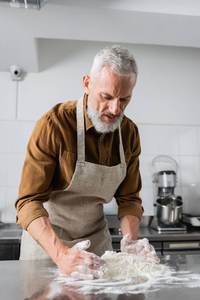 Зрілий бородатий шеф-кухар в фартусі робить тісто на кухонному столі
  - Фото, зображення