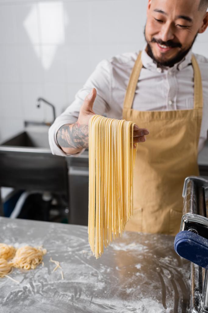 Bulanık Asyalı şef un ve makarna makinesinin yanında çiğ spagetti tutuyor.  - Fotoğraf, Görsel
