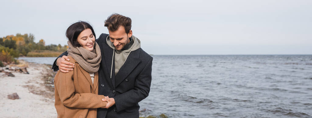 glückliches junges Paar, das sich umarmt und Händchen hält, während es an der Küste spaziert, Banner - Foto, Bild