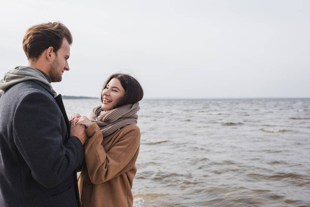 alegre pareja cogida de la mano y mirándose mientras camina cerca del mar - Foto, imagen