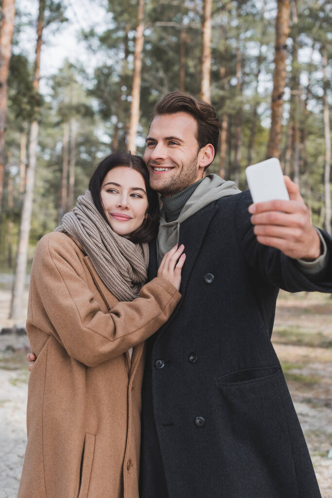 mulher alegre abraçando namorado tomando selfie no telefone celular na floresta de outono - Foto, Imagem