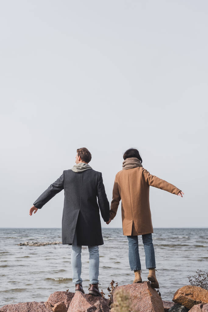Вид сзади молодой пары в осенних пальто, держащейся за руки, стоя на берегу моря - Фото, изображение