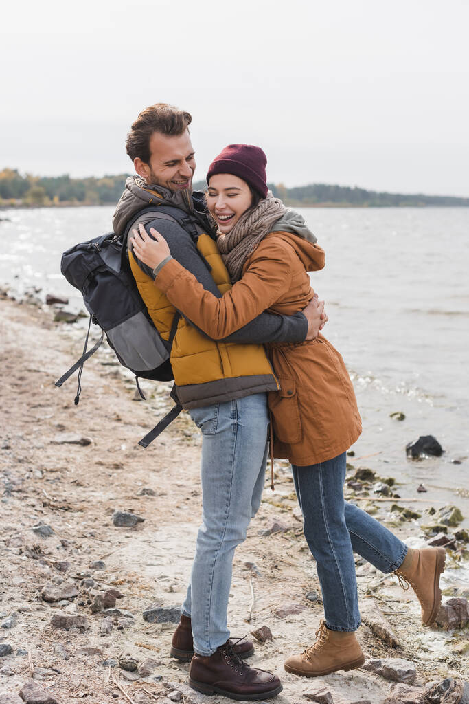 joyful couple in autumn outfit hugging while walking on sea coast - Photo, Image