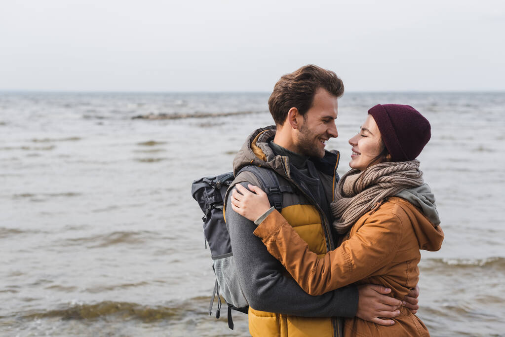 pareja joven en traje de otoño abrazándose y mirándose cerca del mar - Foto, imagen