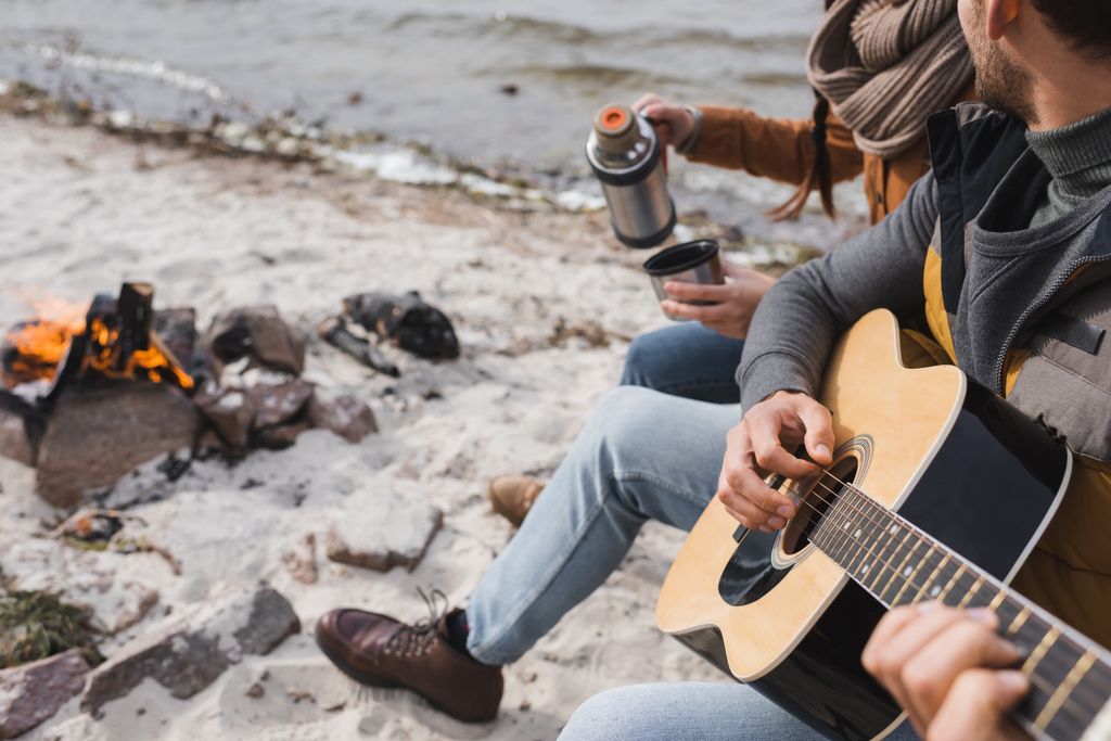 vista parcial del hombre tocando la guitarra cerca de la mujer con termos a orillas del río - Foto, Imagen