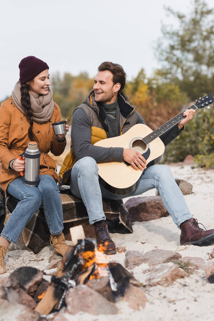 nő termosszal és férfi akusztikus gitár ül kövek közelében tábortűz - Fotó, kép