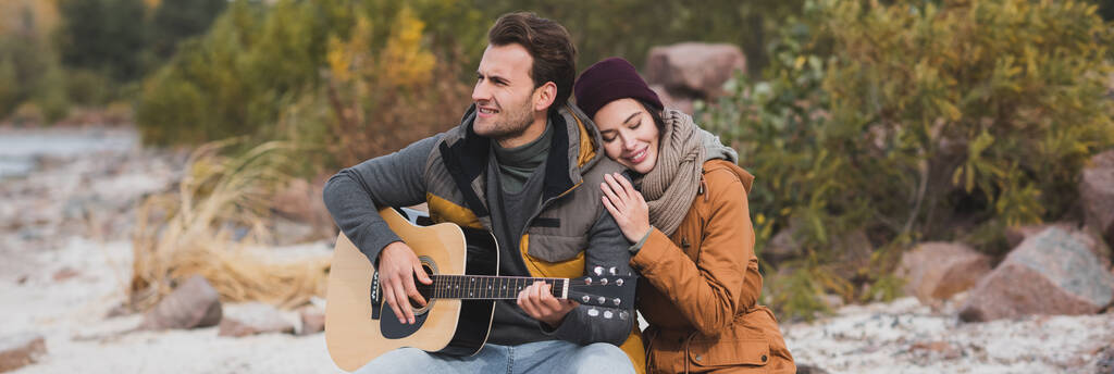 freudige Frau mit geschlossenen Augen umarmt Mann, der draußen Gitarre spielt, Banner - Foto, Bild