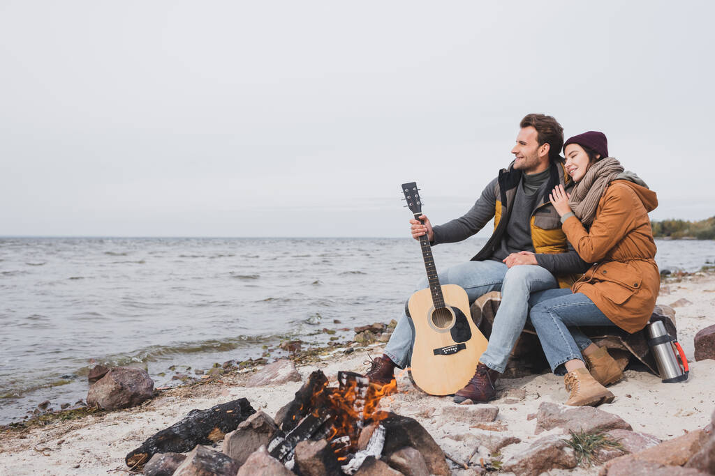 feliz casal com guitarra e thermos sentado perto de fogueira na costa do mar durante parada na caminhada - Foto, Imagem