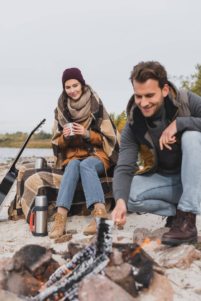 young man near bonfire, and woman sitting with thermo cup under plaid blanket - Photo, Image