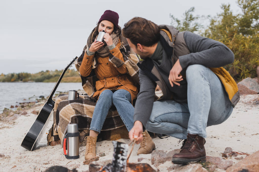 nuori nainen juo termo cup lähellä poikaystävä ja kokko joen rannikolla - Valokuva, kuva