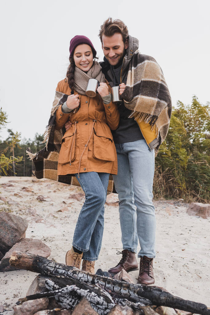 Lächelnde Touristen im herbstlichen Outfit stehen mit Thermobechern am Lagerfeuer - Foto, Bild