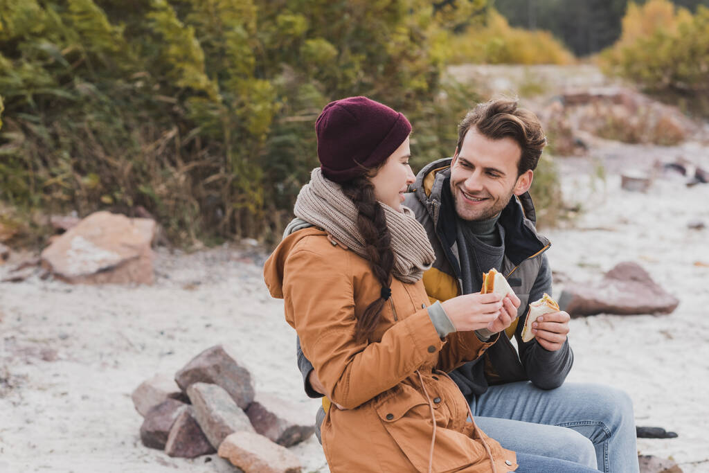 coppia allegra in abiti autunnali parlando e mangiando panini durante la passeggiata all'aperto - Foto, immagini