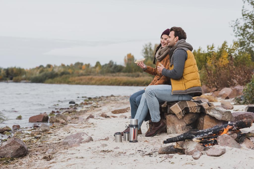 ευτυχισμένο ζευγάρι σε φθινοπωρινά ρούχα κοιτάζοντας το ποτάμι, ενώ κάθεται με σάντουιτς - Φωτογραφία, εικόνα