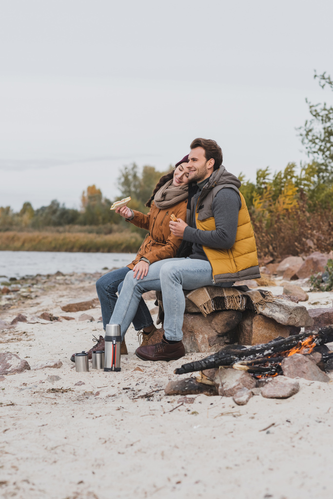 couple heureux avec des sandwichs assis sur des pierres et une couverture près du thermos et du feu de joie - Photo, image