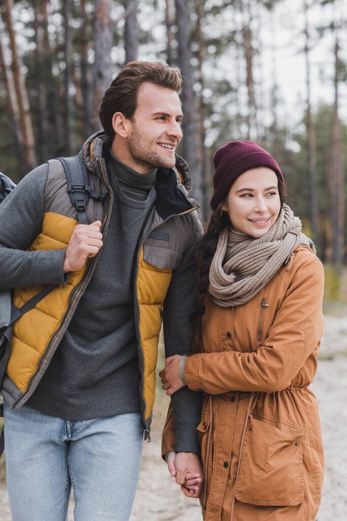 happy young couple in autumn outfit looking away while walking in forest - Photo, Image