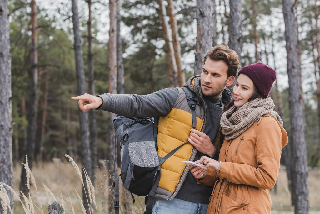 hombre señalando con el dedo cerca de la mujer con el teléfono móvil mientras camina en el bosque - Foto, Imagen