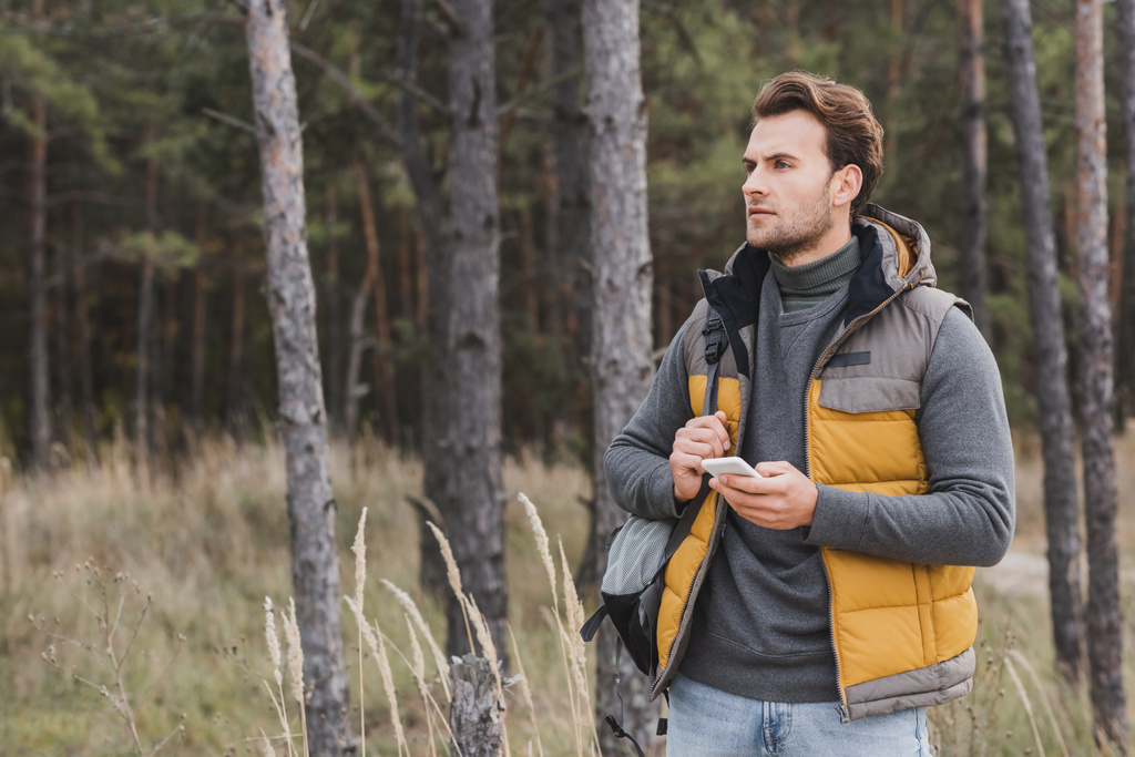 viajero joven con mochila sosteniendo el teléfono móvil mientras camina en el bosque - Foto, imagen