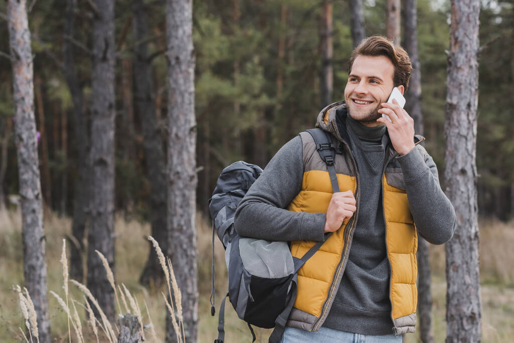 hombre alegre en traje de otoño hablando por teléfono móvil mientras camina en el bosque con mochila - Foto, imagen