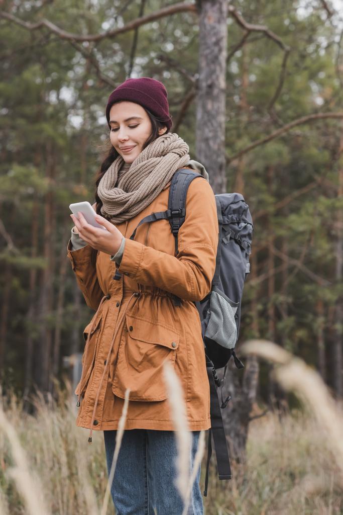 Positive Frau im Herbst-Outfit mit Smartphone auf Richtungssuche im Wald - Foto, Bild