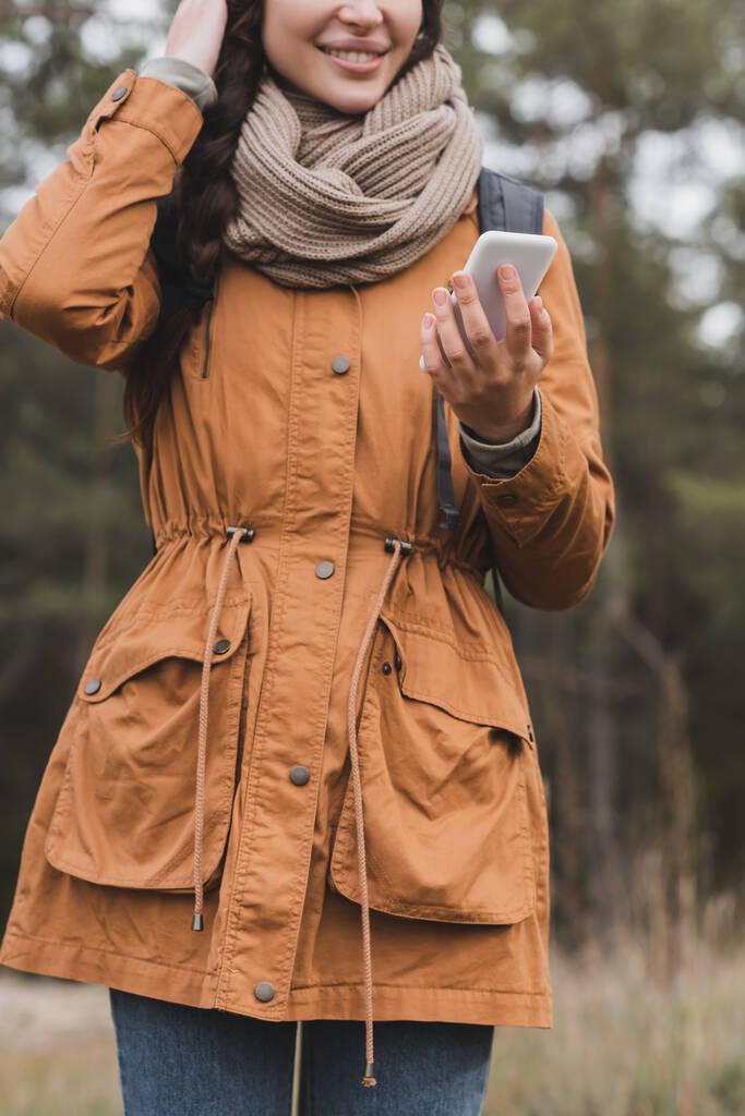 rajattu näkymä hymyilevä nainen syksyllä vaatteita tilalla matkapuhelin aikana kävellä metsässä - Valokuva, kuva