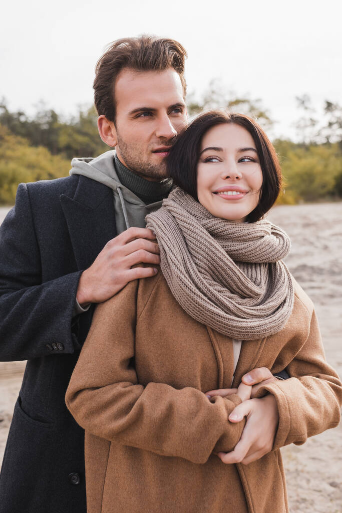 young man hugging cheerful woman in coat and scarf during autumn walk - Photo, Image