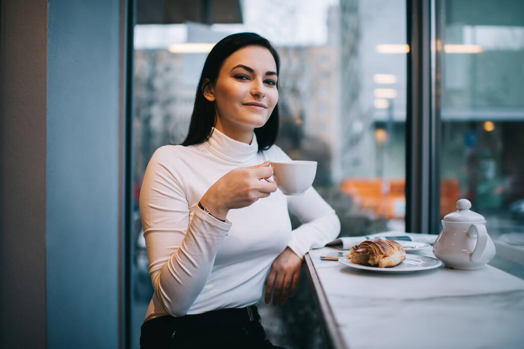 休憩中においしいコーヒーを飲みながらカフェでテーブルに座っている間にカメラを見てカジュアルな服で楽観的な女性 - 写真・画像