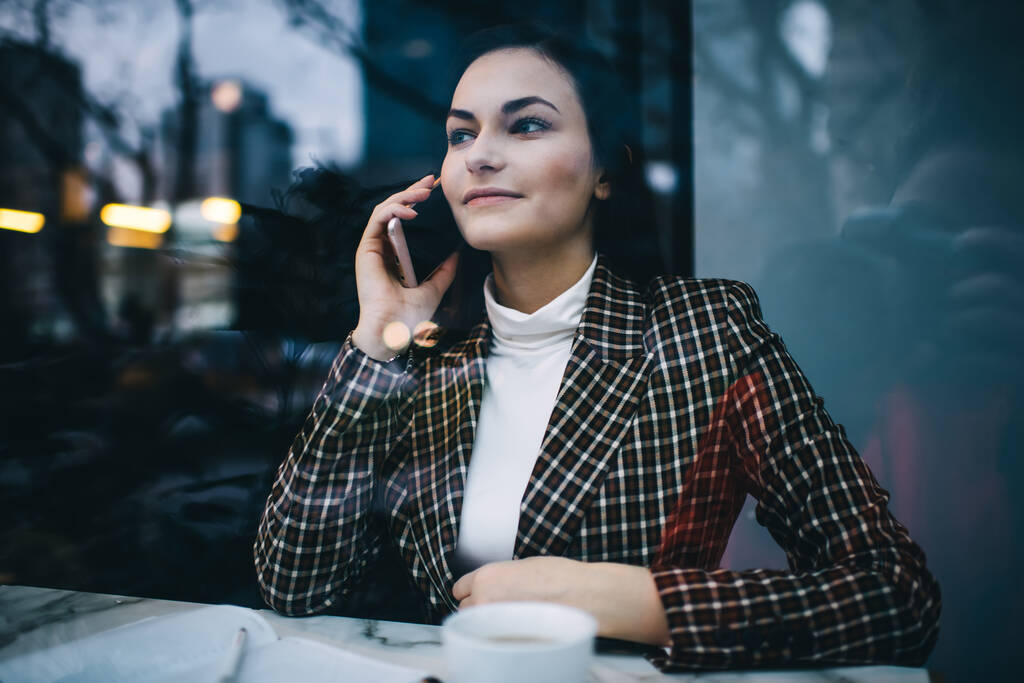 Через окно приятной женщины удаленного работника в модном наряде глядя вдаль звоня на смартфон, сидя за столом в кафе - Фото, изображение