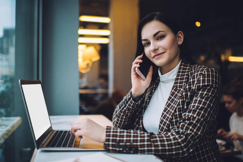 Hymyilevä nuori naispuolinen työntekijä trendikkäässä asussa puhuu matkapuhelimella ja surffaa internetissä kannettavalla tietokoneella työskennellen etänä kahvilassa - Valokuva, kuva