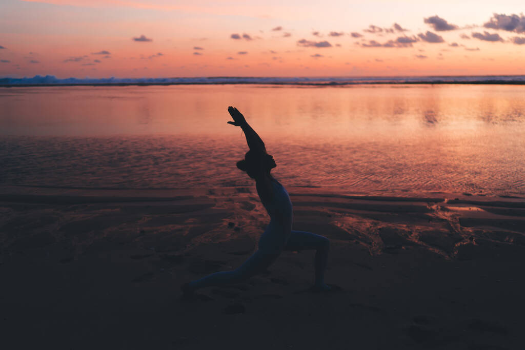 Вид сбоку на гибкий силуэт спортсменки, стоящей в полумесяце Выпад позировать на пустом песчаном пляже против красочного заката неба и спокойной воды во время тренировки - Фото, изображение