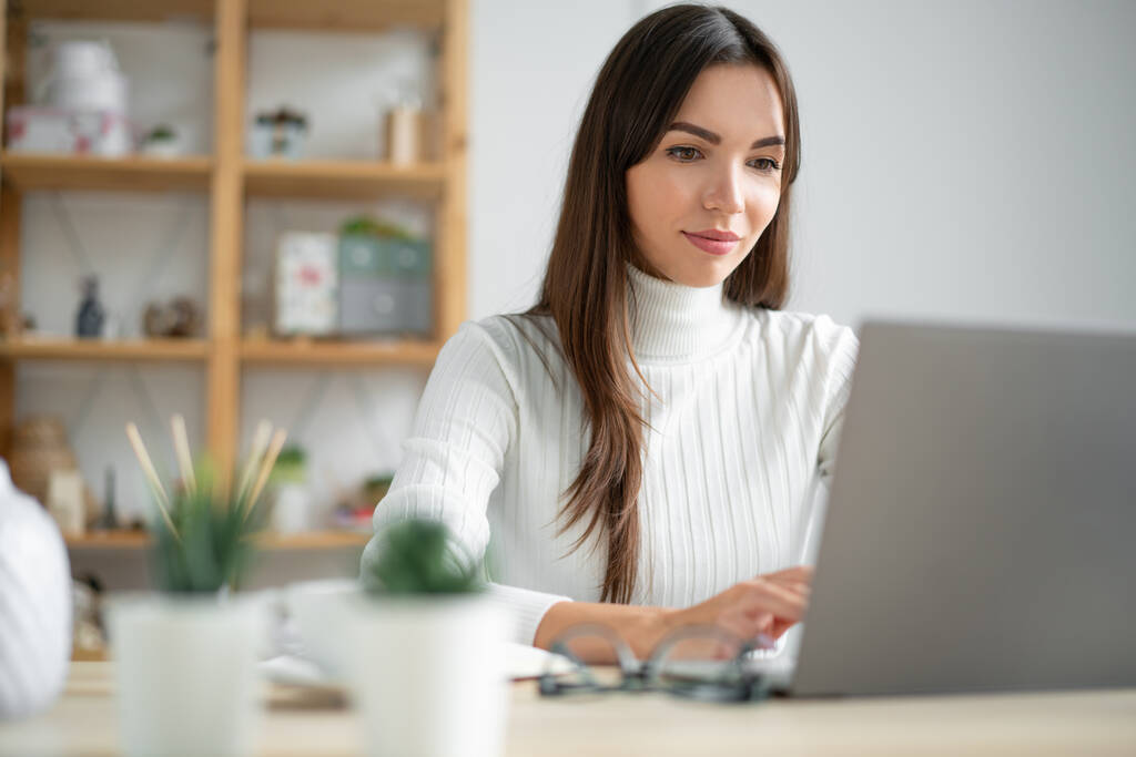 白いセーターを着た若い女性がノートパソコンの後ろで働いています。. - 写真・画像