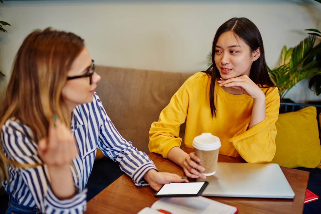Vrolijke Aziatische student in heldere kleren met afhaaldrankje praten met een vriend met behulp van smartphone, terwijl het doorbrengen van tijd in cafe overdag - Foto, afbeelding