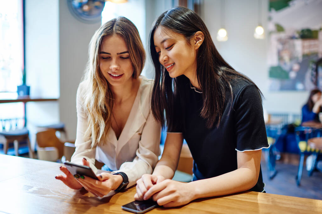 Szczęśliwe przyjaciółki w luźnych ubraniach z zębate uśmiechy siedzi przy stole w kawiarni i przeglądania telefonu komórkowego podczas przerwy - Zdjęcie, obraz