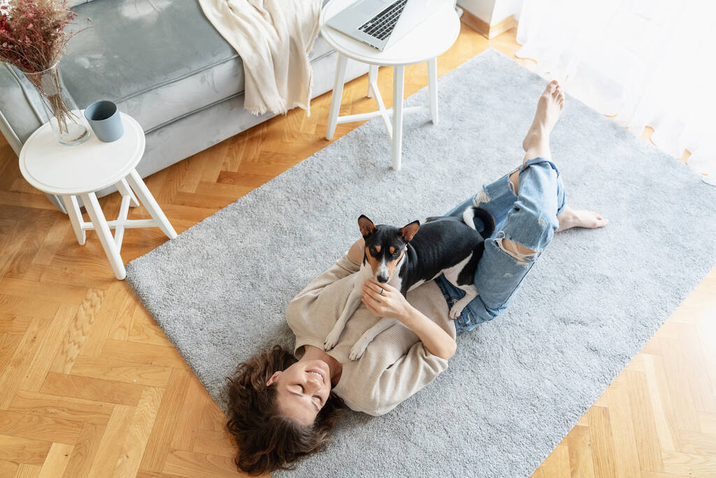 Junge glückliche Frau, die zu Hause mit ihrem Basenji-Hund auf dem Teppich liegt. Hochwertiges Foto - Foto, Bild