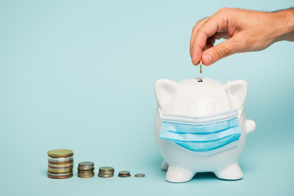 bijgesneden beeld van de mens zetten munt in spaarvarken bank in medisch masker op blauwe, anti-corruptie concept - Foto, afbeelding