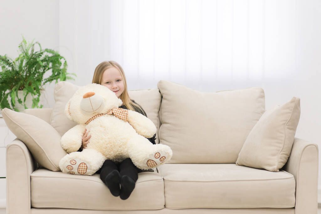 Kleine blonde Mädchen sitzt auf dem Sofa und umarmt ihren Teddybär Foto. - Foto, Bild