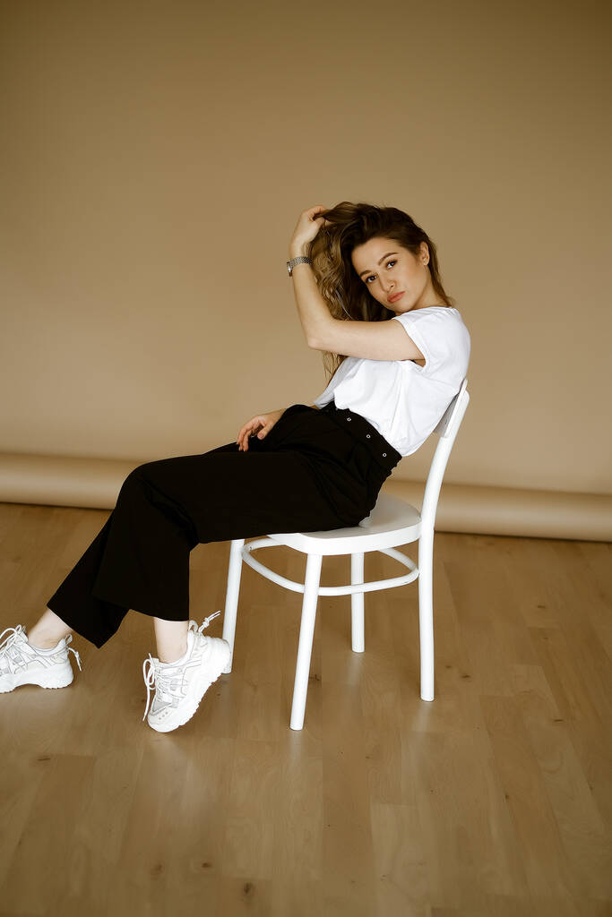 young caucasian woman in black pants and a white T-shirt sitting on a chair, portrait of a brown-eyed brunette with makeup and curly hair, lady in monochrome clothes on a light background - Photo, Image