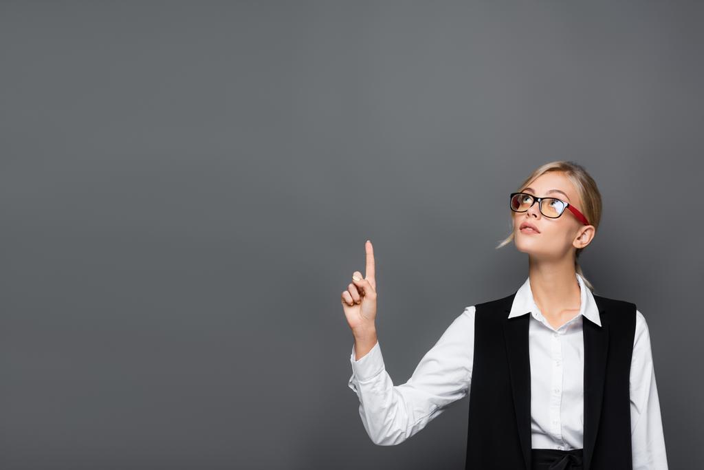 Blonde businesswoman in eyeglasses pointing with finger and looking up isolated on grey  - Photo, Image
