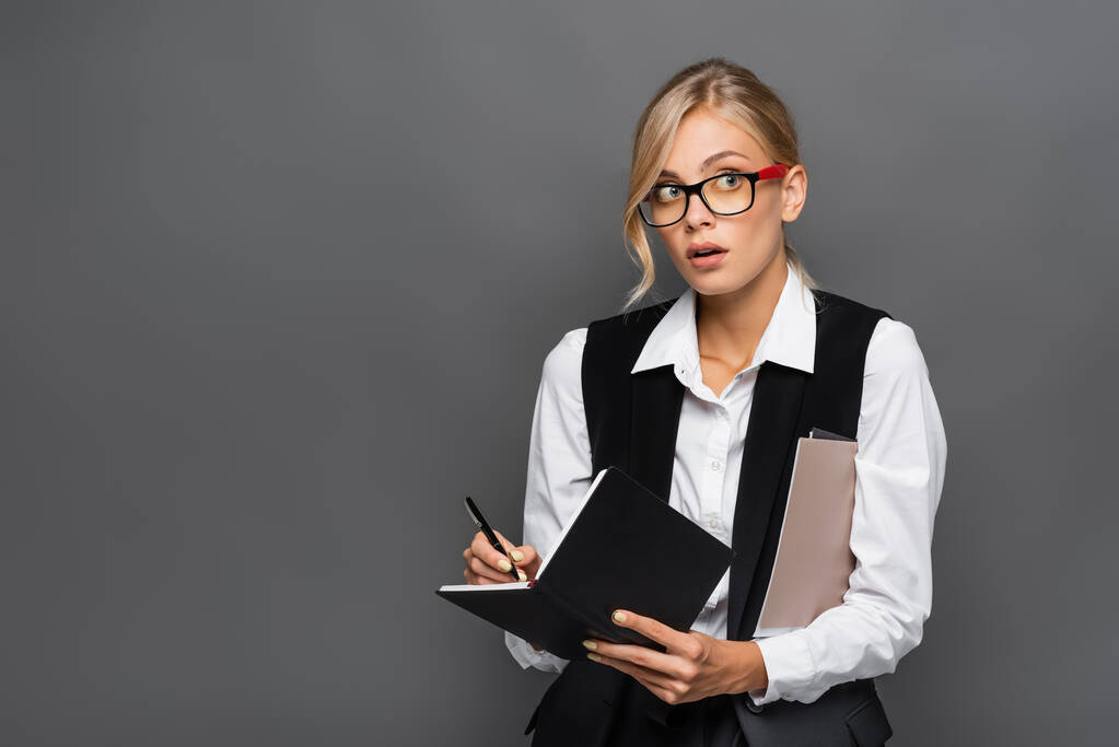 Young businesswoman in eyeglasses writing on notebook and holding paper folders isolated on grey  - Photo, Image
