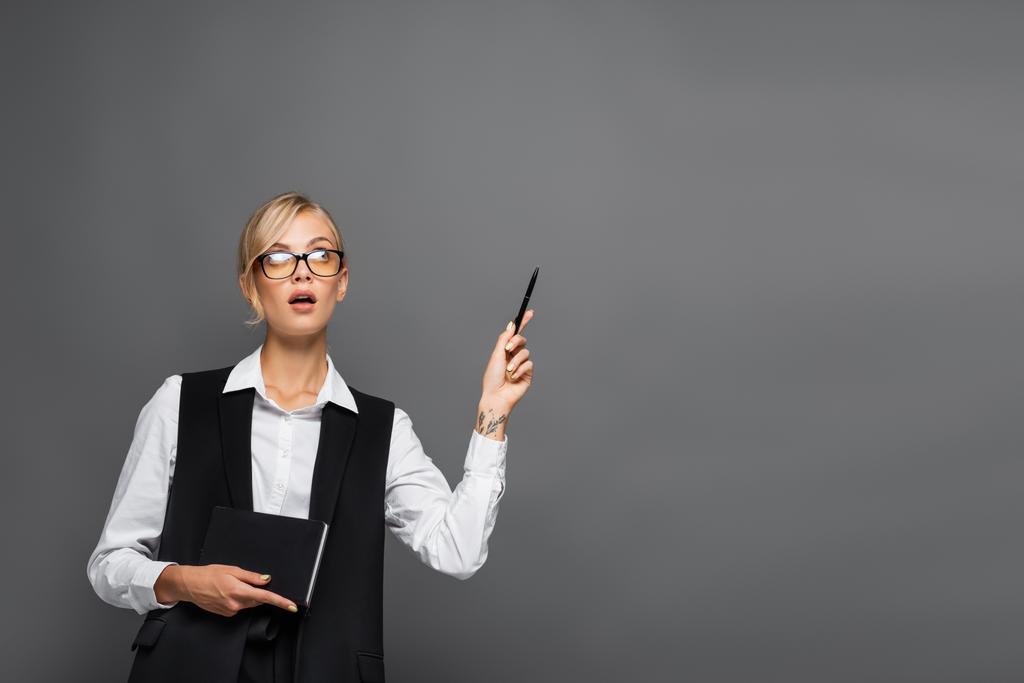 Empresaria pensativa sosteniendo cuaderno y señalando con la mano aislada en gris  - Foto, Imagen