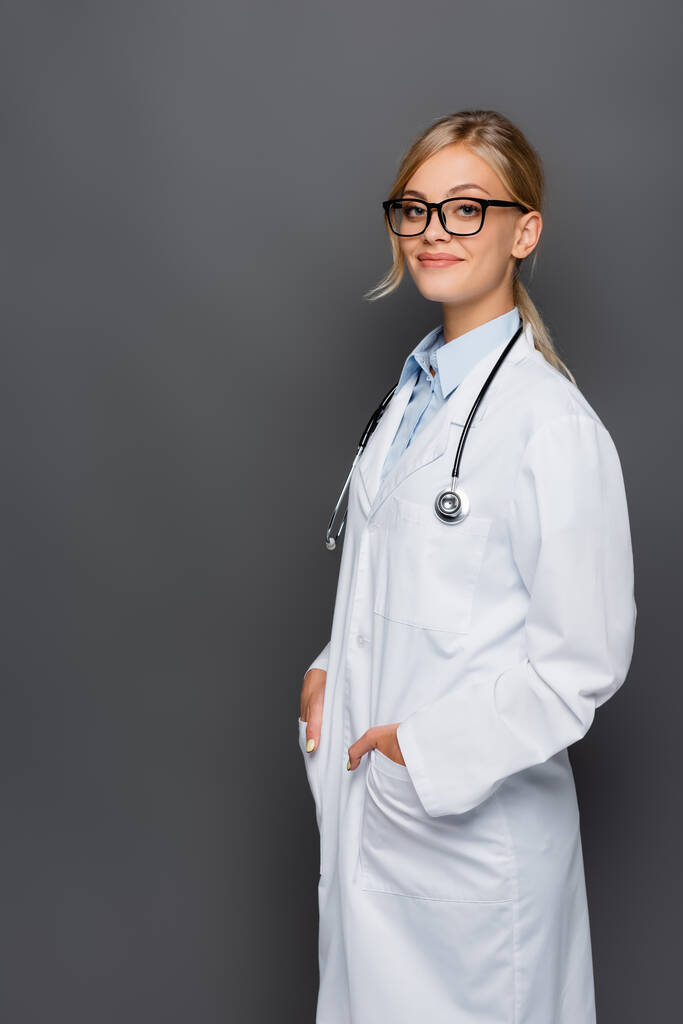 Fröhlicher Arzt in weißem Mantel, die Hände in den Taschen und Blick in die Kamera isoliert auf grau  - Foto, Bild