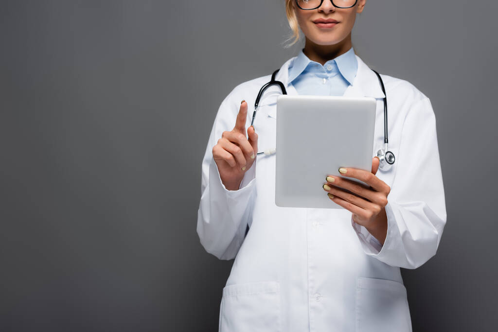 Обрезанный вид молодого врача в белом халате с помощью цифровой таблетки, изолированной на сером  - Фото, изображение