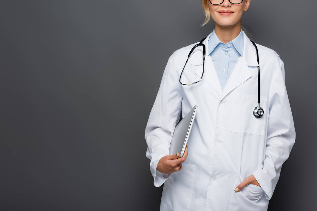 Vista recortada de médico sorridente segurando laptop e mão no bolso de casaco branco isolado em cinza  - Foto, Imagem