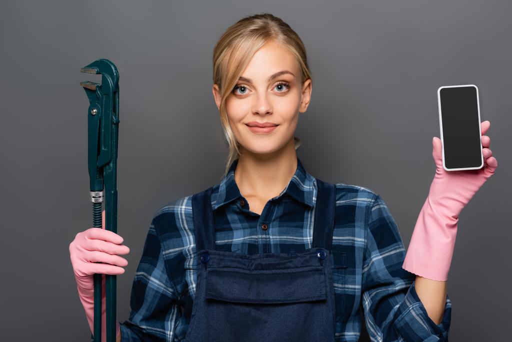 Blonde plumber in overalls holding cellphone with blank screen and pipe wrench isolated on grey  - Photo, Image