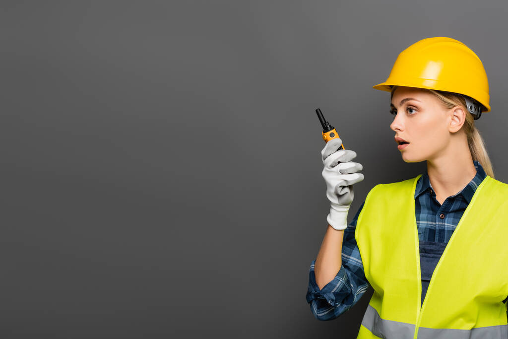 Blondine bouwer in handschoen en veiligheidsvest met walkietalkie geïsoleerd op grijs  - Foto, afbeelding