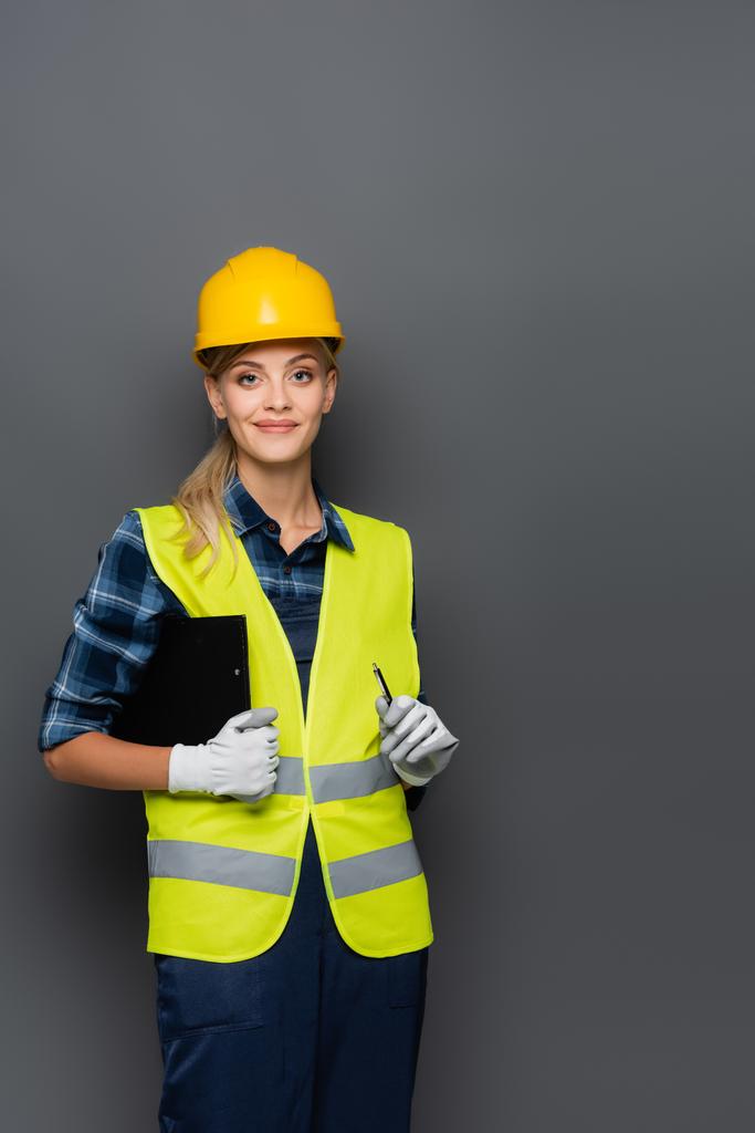 Cheerful builder in hardhat looking at camera while holding clipboard and pen isolated on grey  - Photo, Image