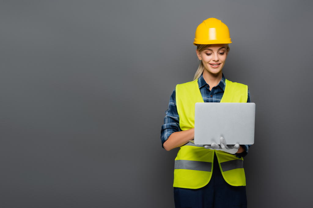 Позитивний будівельник у хардхаті та захисному жилеті з використанням ноутбука ізольовано на сірому
  - Фото, зображення