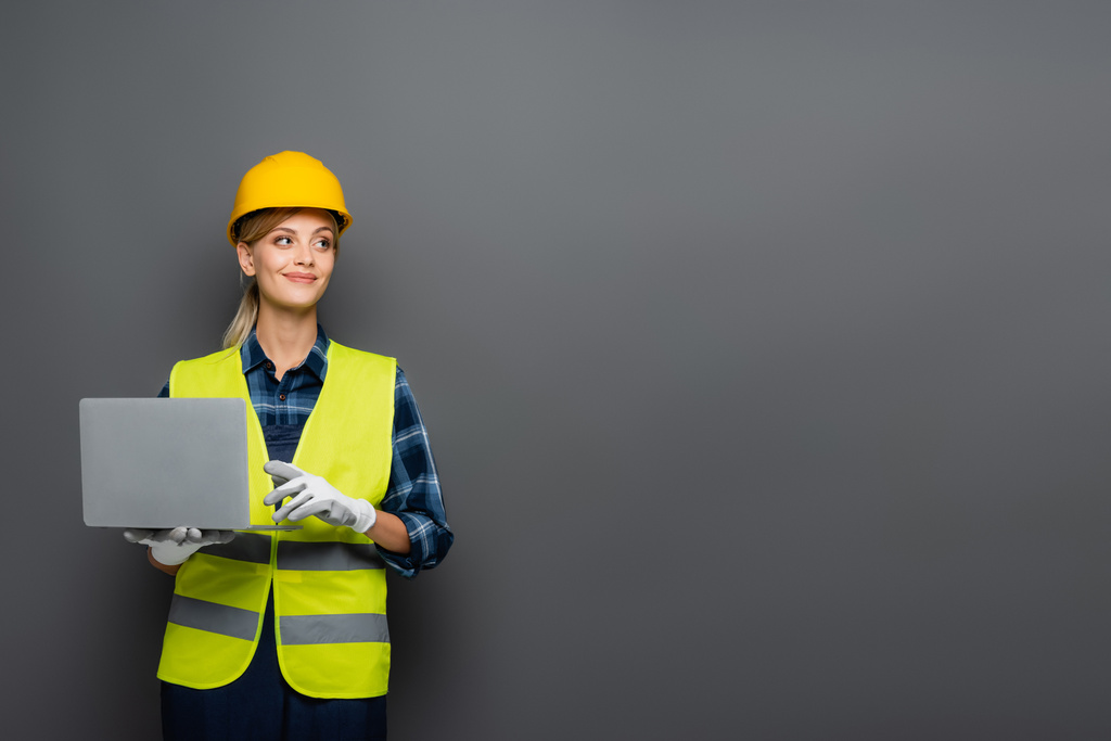 Веселий будівельник в рукавичках і шоломі тримає ноутбук ізольований на сірому
  - Фото, зображення