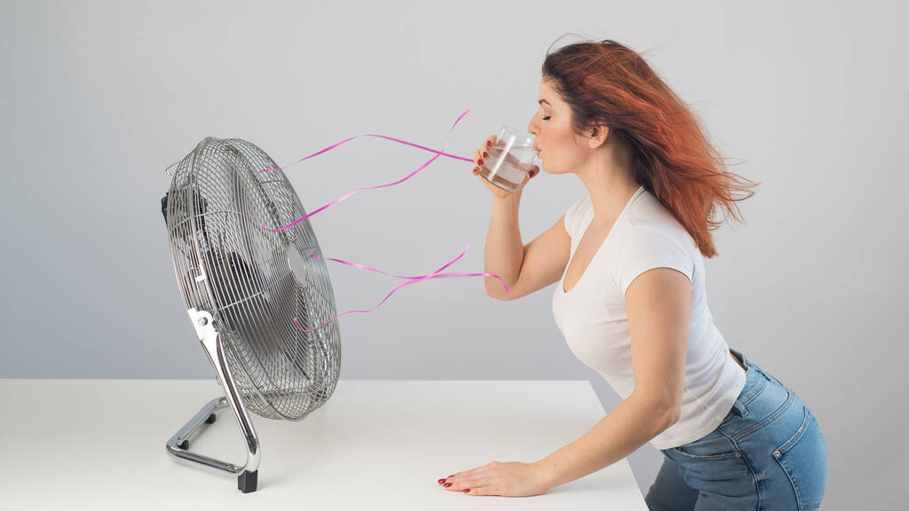 Una mujer caucásica pelirroja se enfría junto al ventilador eléctrico y bebe una bebida fría. Climatización en el apartamento - Foto, imagen