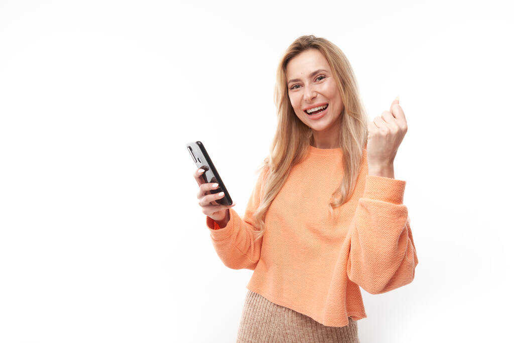 τυχερό χαμογελαστό ξανθό κορίτσι κρατώντας smartphone σε casual απομονωμένο σε λευκό φόντο στούντιο έλαβε χαρούμενο μήνυμα, γιορτάζοντας τη νίκη - Φωτογραφία, εικόνα